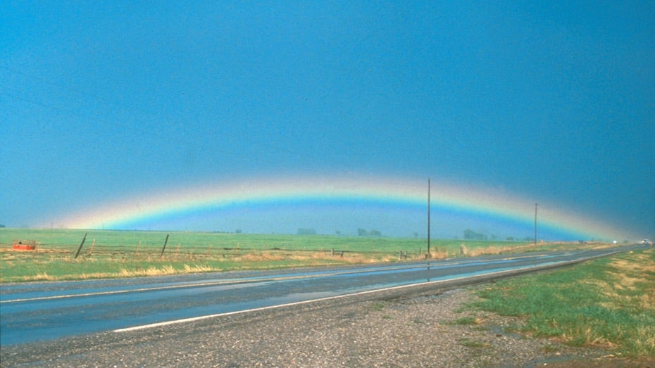 A highway in the plains with a full arc rainbow in the background.